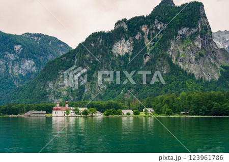 A white church with red domes stands by the tranquil waters of a lake, surrounded by lush greenery and majestic mountains on a sunny day. Konigssee lake Bavaria German Alps Berchtesgaden church by the 123961786