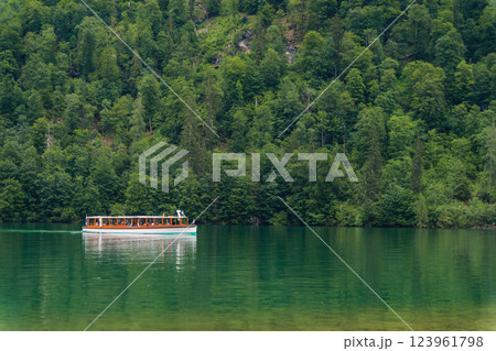 A boat gracefully moves through the calm waters of a beautiful lake, bordered by dramatic green hills and dense forests under a cloudy sky. The peaceful setting invites relaxation. Konigssee Bavaria A boat gracefully moves through the calm waters of a beautiful lake, bordered by dramatic green hills and dense forests under a cloudy sky. The peaceful setting invites relaxation. Konigssee Bavaria 123961798