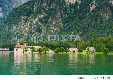 A white church with red domes stands by the tranquil waters of a lake, surrounded by lush greenery and majestic mountains on a sunny day. Konigssee lake Bavaria German Alps Berchtesgaden church by the 123961916