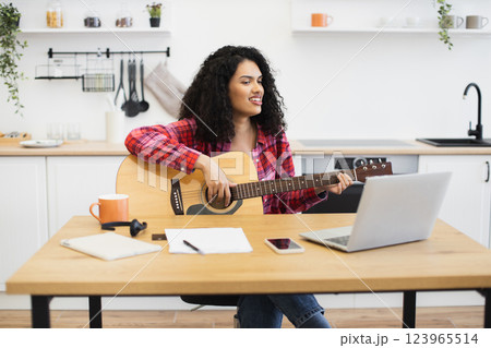Young adult female with curly hair playing guitar in kitchen at table with laptop, illustrating home music practice or creative remote work lifestyle combining leisure and productivity. Young adult female with curly hair playing guitar in kitchen at table with laptop, illustrating home music practice or creative remote work lifestyle combining leisure and productivity. 123965514
