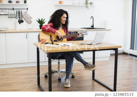 Young adult female with curly hair playing guitar in kitchen at table with laptop, illustrating home music practice or creative remote work lifestyle combining leisure and productivity. 123965515