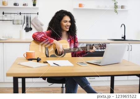 Young adult female with curly hair playing guitar in kitchen at table with laptop, illustrating home music practice or creative remote work lifestyle combining leisure and productivity. 123965522