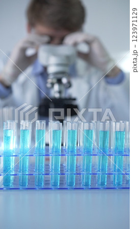 Test tubes with a blue liquid, close-up view. Male scientist wearing white protective gloves and glasses is using microscope on the background. Science and Medicine concept Test tubes with a blue liquid, close-up view. Male scientist wearing white protective gloves and glasses is using microscope on the background. Science and Medicine concept 123971129