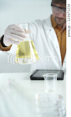 Man scientist wearing white protective gloves, and glasses is holding a yellow chemical solution inside an Erlenmeyer flask in a laboratory, vertical portrait Man scientist wearing white protective gloves, and glasses is holding a yellow chemical solution inside an Erlenmeyer flask in a laboratory, vertical portrait 123971238