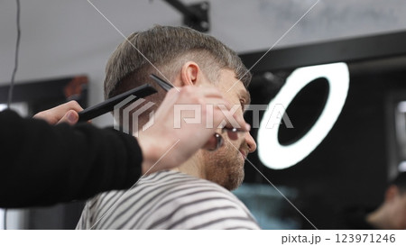 Professional barber trimming blond heard male client's hair with scissors and comb, creating precise modern hairstyle inside contemporary grooming salon Professional barber trimming blond heard male client's hair with scissors and comb, creating precise modern hairstyle inside contemporary grooming salon 123971246
