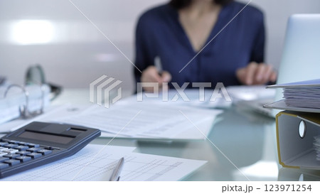 Black calculator, pen and documents are lying on accountant's desk. Businesswoman wearing blue dress is working with documents on the background. Business and audit concept Black calculator, pen and documents are lying on accountant's desk. Businesswoman wearing blue dress is working with documents on the background. Business and audit concept 123971254