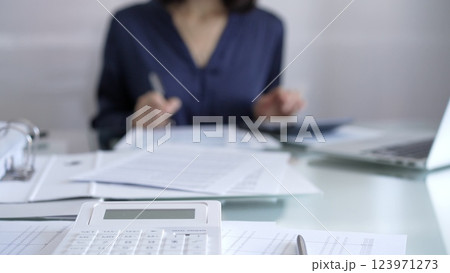 White calculator, pen and documents are lying on accountant's desk. Businesswoman wearing blue dress is working with documents on the background. Business and audit concept White calculator, pen and documents are lying on accountant's desk. Businesswoman wearing blue dress is working with documents on the background. Business and audit concept 123971273