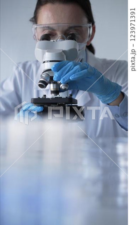 Woman scientist wearing blue gloves, protective glasses and medical mask is using microscope in modern laboratory setup. Low key lighting photo. Science and Medicine Woman scientist wearing blue gloves, protective glasses and medical mask is using microscope in modern laboratory setup. Low key lighting photo. Science and Medicine 123971391