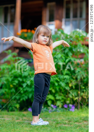 A young girl stands in a green garden with arms outstretched as if balancing or pretending to fly. 123971496