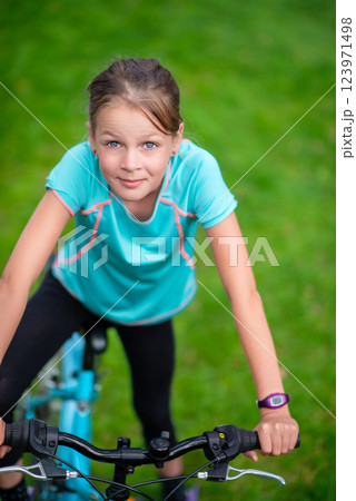 Smiling young girl riding a blue bicycle outdoors in a green park, enjoying an active lifestyle and outdoor fun in summer 123971498