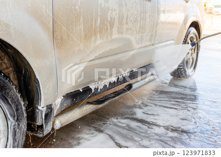 Close-up of a car being washed with high-pressure water and foam at a self-service car wash, focusing on the wheel and side panel covered in soap suds 123971833