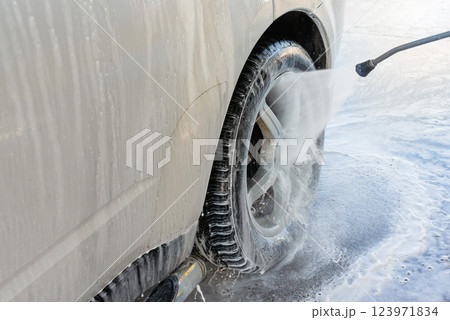 Close-up of a car being washed with high-pressure water and foam at a self-service car wash, focusing on the wheel and side panel covered in soap suds 123971834