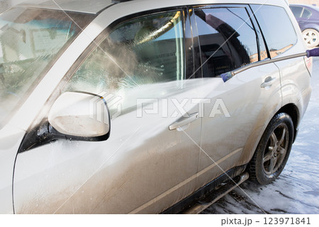 A silver SUV being cleaned with a high-pressure water spray at an outdoor car wash. The image captures the contrast between the dirty and clean surfaces 123971841