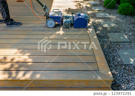 Worker cleaning and sanding a wooden deck with a specialized machine in a backyard setting with lush green lawn and a bright sunny environment. Worker cleaning and sanding a wooden deck with a specialized machine in a backyard setting with lush green lawn and a bright sunny environment. 123972291