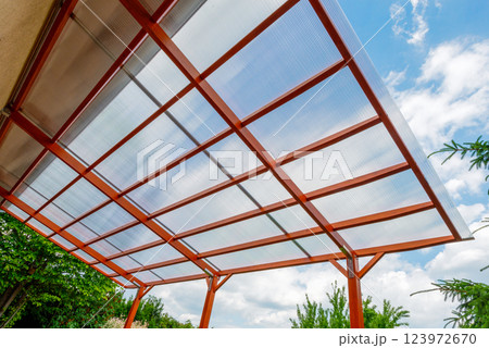 Transparent polycarbonate roof on a modern wooden pergola structure in a backyard, providing shade and protection with a clear sky background. 123972670