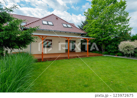 Modern suburban house with a red tiled roof, wooden deck with a transparent pergola, and a beautifully landscaped backyard featuring green grass and lush trees on a sunny day Modern suburban house with a red tiled roof, wooden deck with a transparent pergola, and a beautifully landscaped backyard featuring green grass and lush trees on a sunny day 123972675