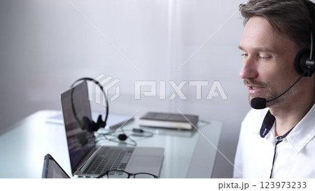 Businessman wearing headset and glasses, using laptop for video conference call, working remotely from home office. Call center concept 123973233