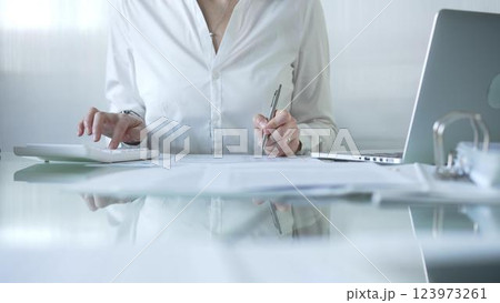 Businesswoman in blue blouse is using white calculator and taking notes while working on financial report in contemporary office setting. Audit and taxes concept Businesswoman in blue blouse is using white calculator and taking notes while working on financial report in contemporary office setting. Audit and taxes concept 123973261
