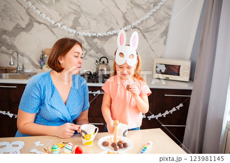 Mid shot of little girl in paper rabbit-mask and her mother drink tea and eat sweets together, sitting at a festive Easter table. Spring holiday 123981145