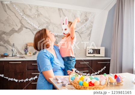 Wide shot of Caucasian happy mother and her child are hugging nearby festive table for Easter. A cute little girl in a paper rabbit mask Wide shot of Caucasian happy mother and her child are hugging nearby festive table for Easter. A cute little girl in a paper rabbit mask 123981165