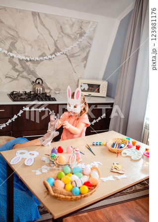 Wide shot of young mother plays with rabbit glove with her little daughter in a decorated kitchen. Funny girl in a paper rabbit mask is having fun with her mother. Vertical 123981176