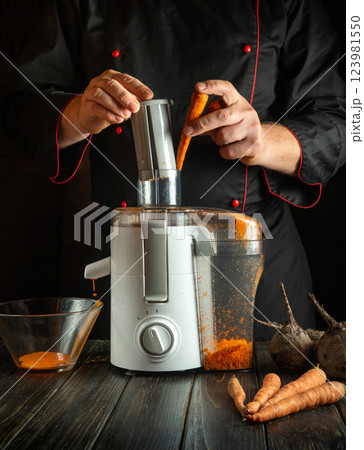 Preparation of vegetable drink with carrots and beets on an electric mixer. Work environment on the kitchen table. The chef adds fresh carrots to the juicer with his hands 123981550