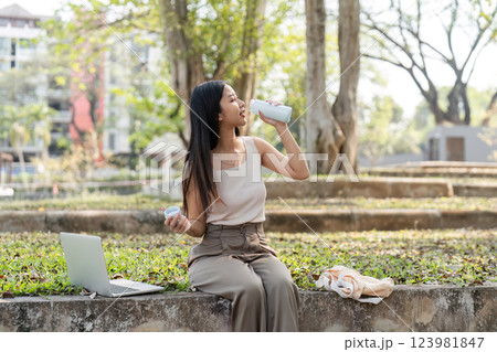 Sustainable lifestyle. Young woman enjoying a refreshing drink while working on her laptop outdoors. Sustainable lifestyle. Young woman enjoying a refreshing drink while working on her laptop outdoors. 123981847