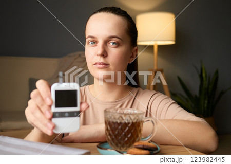 A person is holding a glucometer while sitting at a table with a cup of tea and cookies. The setting is a cozy indoor area, indicating a moment of health monitoring 123982445