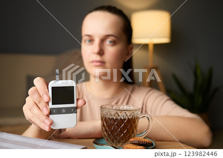 In a cozy indoor setting, a woman checks her blood sugar levels with a glucometer. She holds the device in one hand while a cup of tea and cookies are on the table nearby 123982446
