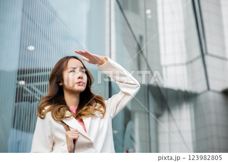 Happy woman wearing white suit jacket with brown bag at sunlight outdoors. Portrait of confidence young businesswoman standing outside office building in city raise your hand to shade sun 123982805