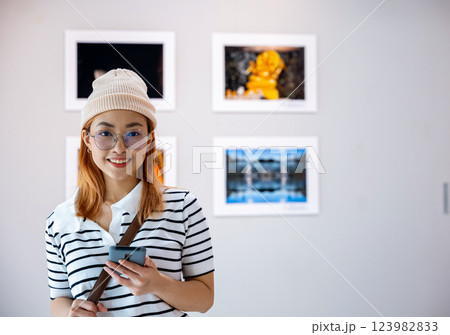 Asian female watching at photo frame with smartphone at artwork gallery show, Visitor woman smiling at picture art gallery collection in front framed paintings pictures she holding mobile phone 123982833