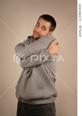 A young man with a light beard gently hugs himself with both arms, creating a cozy and emotional vibe. His gesture expresses self-love and warmth. The scene feels intimate and comforting. 123985444