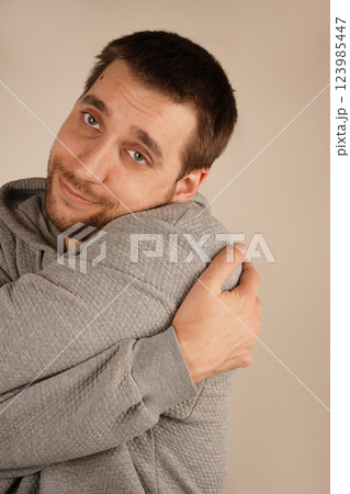 His gesture expresses self-love and warmth. The scene feels intimate and comforting. A young man with a light beard gently hugs himself with both arms, creating a cozy and emotional vibe. 123985447