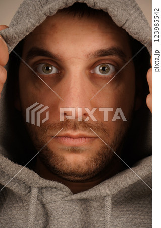 His relaxed posture and urban style suggest a sense of casual coolness. The hoodie creates a street-style vibe. A young man in a grey hoodie with a hood pulls it over with both hands. 123985542