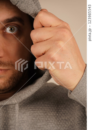 The hoodie creates a street-style vibe. A young man in a grey hoodie with a hood pulls it over with both hands. His relaxed posture and urban style suggest a sense of casual coolness. The hoodie creates a street-style vibe. A young man in a grey hoodie with a hood pulls it over with both hands. His relaxed posture and urban style suggest a sense of casual coolness. 123985543