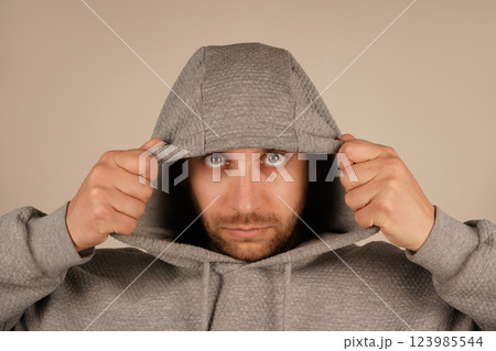 A young man in a grey hoodie with a hood pulls it over with both hands. His relaxed posture and urban style suggest a sense of casual coolness. The hoodie creates a street-style vibe. A young man in a grey hoodie with a hood pulls it over with both hands. His relaxed posture and urban style suggest a sense of casual coolness. The hoodie creates a street-style vibe. 123985544