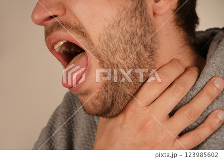 His facial expression conveys discomfort and a possible choking sensation. A man holds his throat with one hand, opening his mouth as if he is coughing or struggling to breathe. His facial expression conveys discomfort and a possible choking sensation. A man holds his throat with one hand, opening his mouth as if he is coughing or struggling to breathe. 123985602