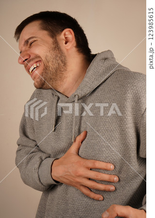 A man in a gray hoodie holds his chest near the ribs with one hand, his face showing pain. The expression suggests a burning sensation or severe discomfort. It could indicate heart failure symptoms. A man in a gray hoodie holds his chest near the ribs with one hand, his face showing pain. The expression suggests a burning sensation or severe discomfort. It could indicate heart failure symptoms. 123985615