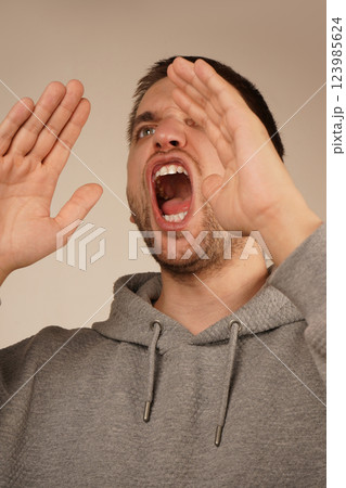 His actions suggest a loud announcement. The image conveys excitement and energy. With dark hair and a lively expression, the man cups both hands over his mouth, resembling a megaphone. His actions suggest a loud announcement. The image conveys excitement and energy. With dark hair and a lively expression, the man cups both hands over his mouth, resembling a megaphone. 123985624