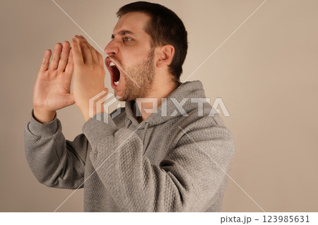 His actions suggest a loud announcement. The image conveys excitement and energy. With dark hair and a lively expression, the man cups both hands over his mouth, resembling a megaphone. His actions suggest a loud announcement. The image conveys excitement and energy. With dark hair and a lively expression, the man cups both hands over his mouth, resembling a megaphone. 123985631