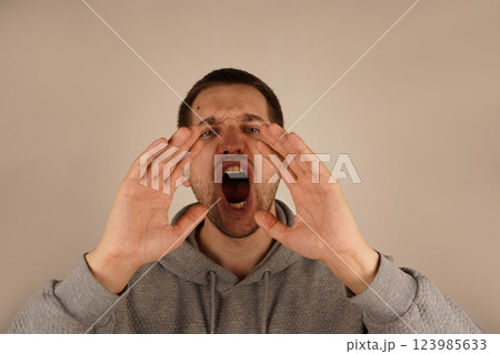 The dark-haired man holds both palms around his mouth, forming a makeshift megaphone. His animated gesture implies shouting or announcing something important. His expression radiates urgency. The dark-haired man holds both palms around his mouth, forming a makeshift megaphone. His animated gesture implies shouting or announcing something important. His expression radiates urgency. 123985633