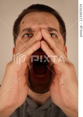 The scene captures his dramatic gesture. A young man with dark hair places his hands near his mouth, resembling a megaphone. His enthusiastic pose suggests he is delivering an energetic shout. The scene captures his dramatic gesture. A young man with dark hair places his hands near his mouth, resembling a megaphone. His enthusiastic pose suggests he is delivering an energetic shout. 123985634