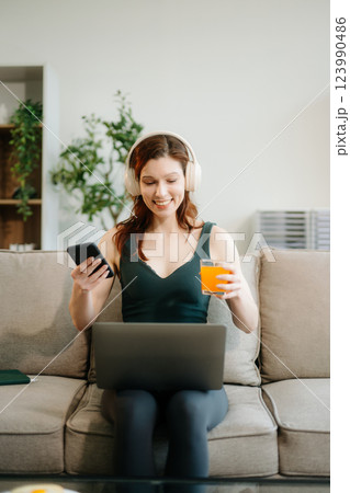 Happy woman with headphones using a laptop in a cozy living room with food and drink. Perfect for themes Happy woman with headphones using a laptop in a cozy living room with food and drink. Perfect for themes 123990486