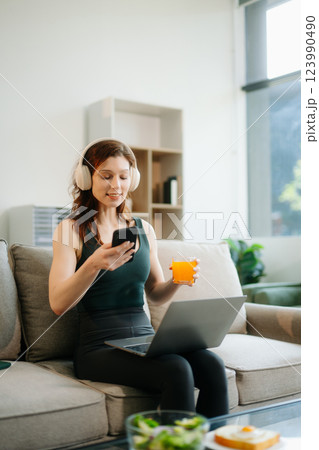 Happy woman with headphones using a laptop in a cozy living room with food and drink. Perfect for themes Happy woman with headphones using a laptop in a cozy living room with food and drink. Perfect for themes 123990490