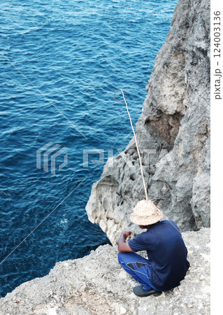An elderly Balinese man wearing straw hat and fishing on the edge of a sea cliff 124003136
