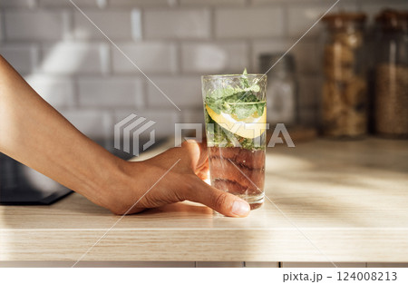 Close-up of female hand holds a refreshing lemon mojito on a wooden table on a kitchen at the home. 124008213