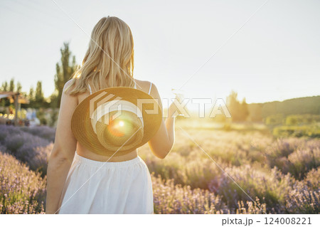 Young female blonde in a white dress and a straw hat holds a glass of wine in a lavender field. Young female blonde in a white dress and a straw hat holds a glass of wine in a lavender field. 124008221