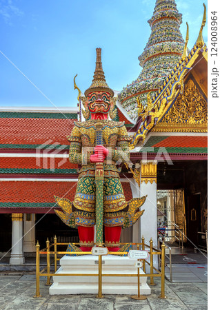 Majestic Yaksha guardian statue with red skin and intricate golden armor stands in front of an ornate Wat Phra Kaew, symbolizing protection and mythology in Thai culture. 124008964