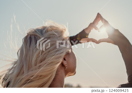 Sun Heart Hands Woman - A woman with long blonde hair holds her hands up to the sun in a heart shape. 124009425