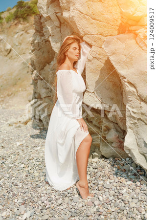 A woman in a white dress is standing on a rocky beach. Concept of serenity and tranquility, as the woman is enjoying the natural beauty of the beach. 124009517